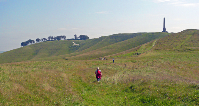 people walking up a steep grassy hill with a carving of a white horse on the hillside